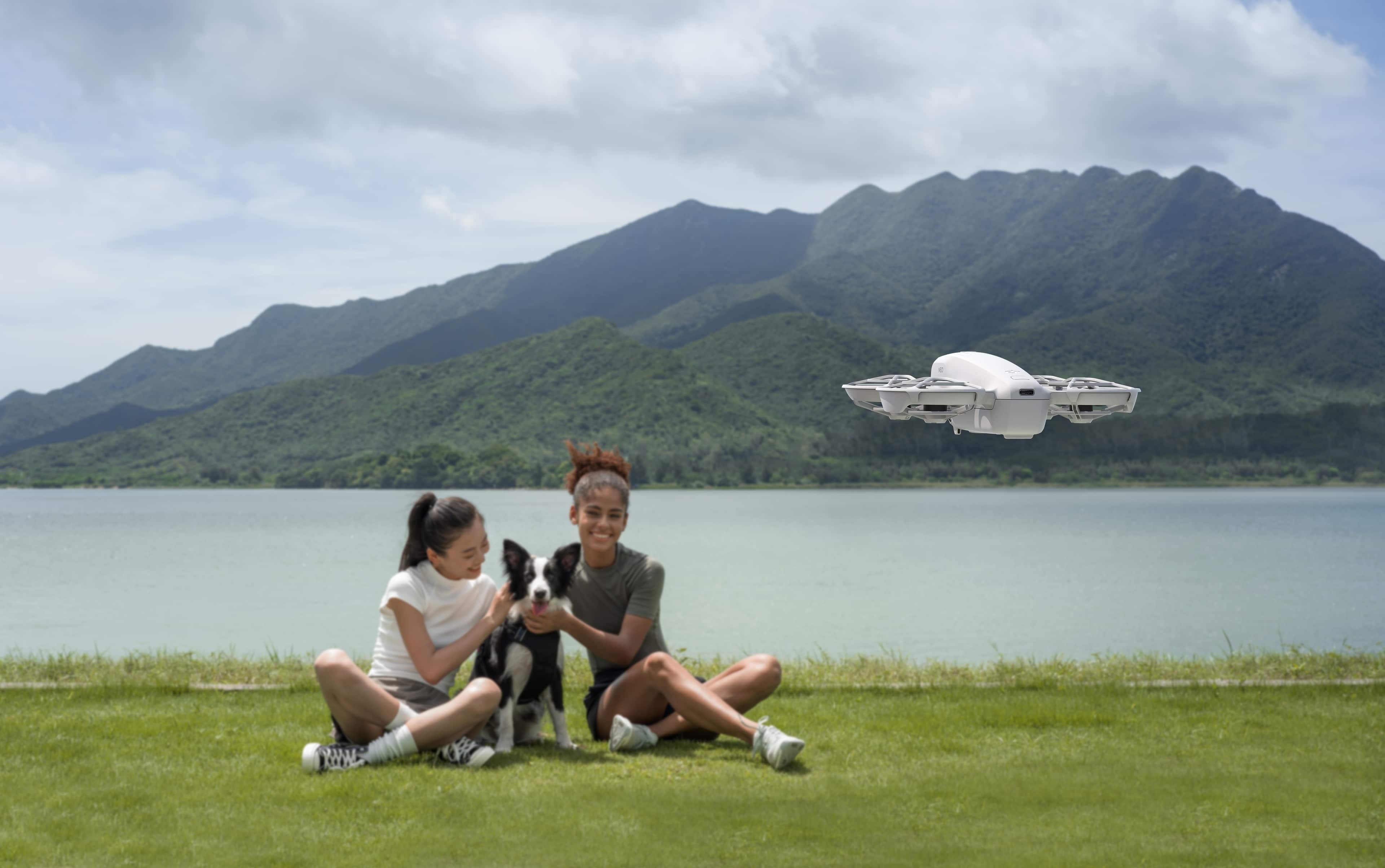 DJI Neo Drohne fliegt über zwei Mädchen mit Hund am Ufer eines Sees, umgeben von Bergen.
