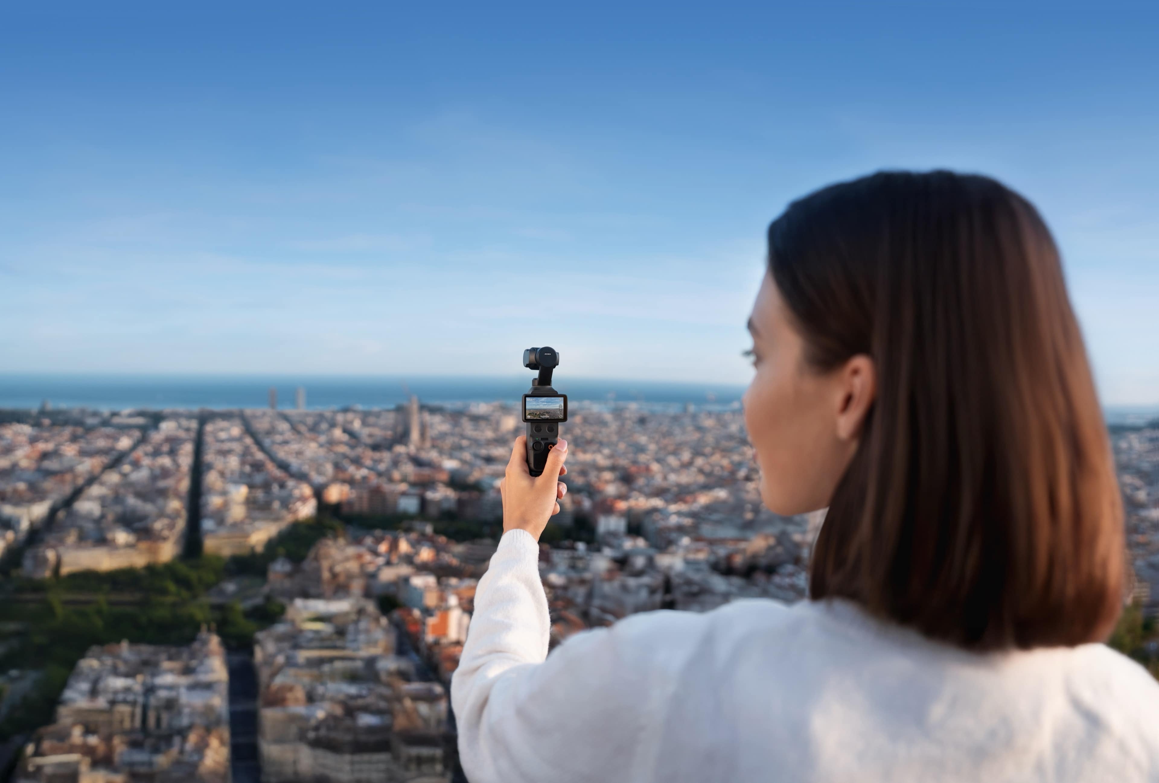 Frau hält die DJI Osmo Pocket 4 Creator Combo und blickt auf eine Stadtlandschaft mit Meer im Hintergrund.