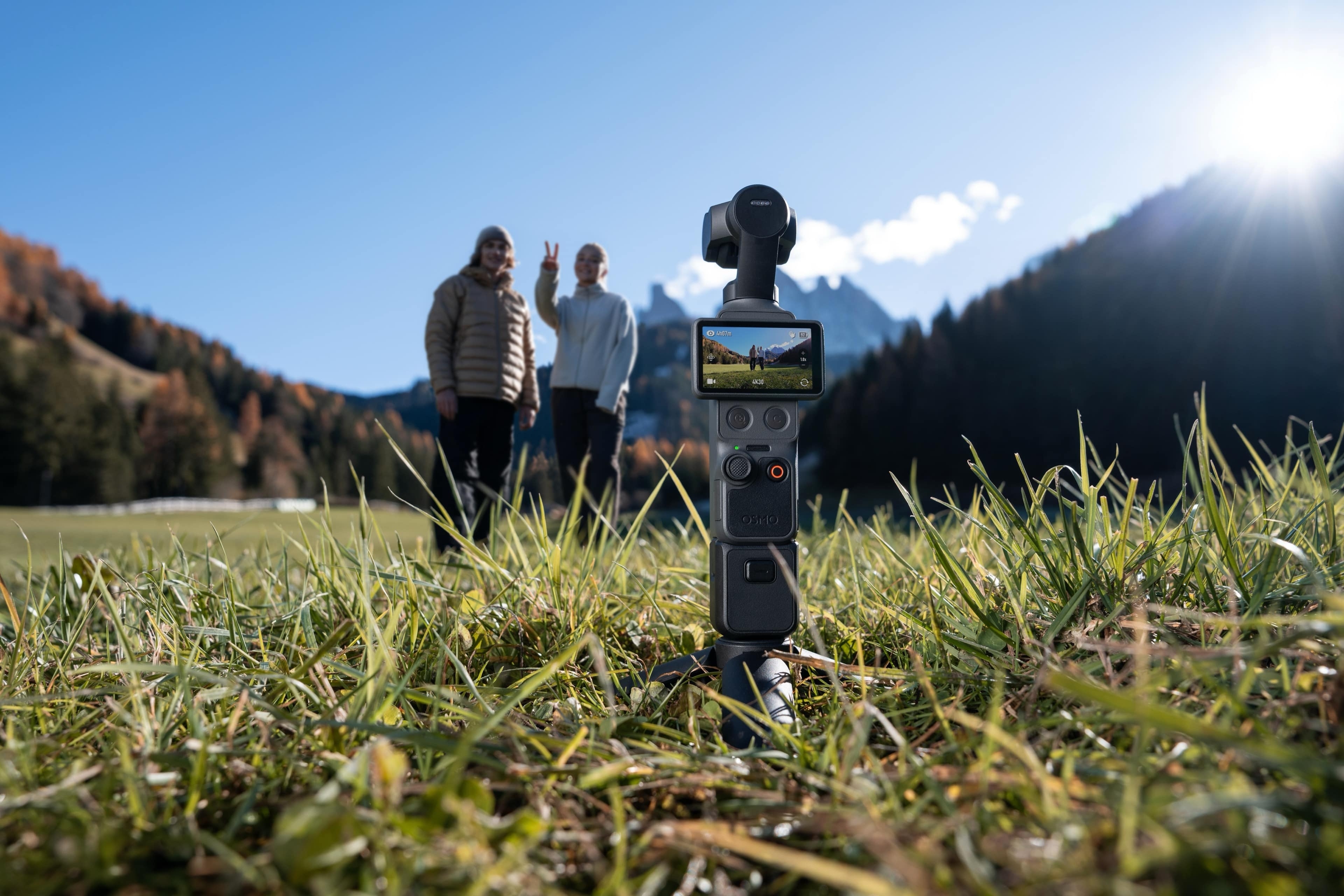 DJI Osmo Pocket 4 Creator Combo auf grassigem Boden, mit zwei Personen im Hintergrund, die winken.