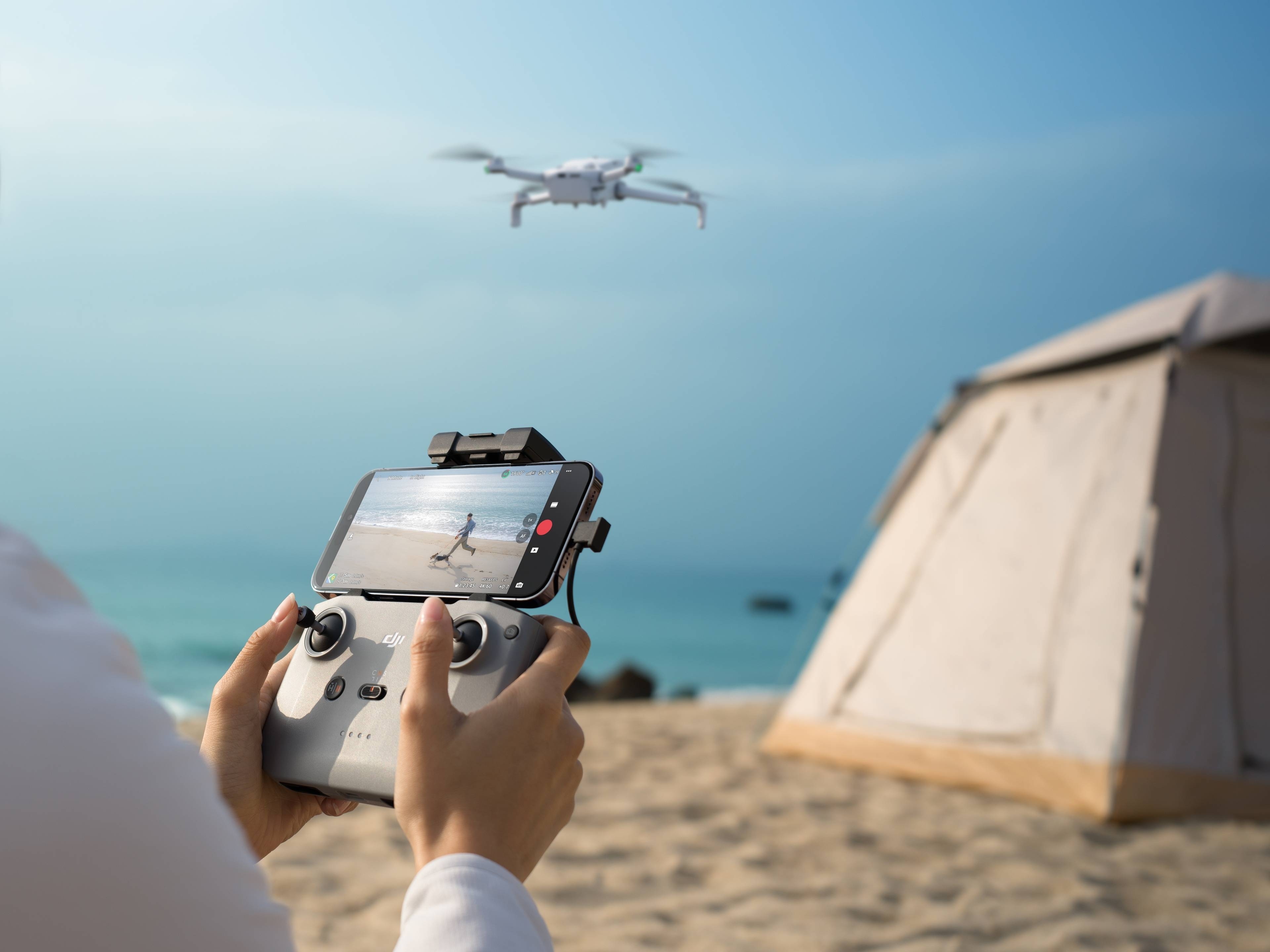 Person steuert die DJI Lito 1 Drohne am Strand mit Fernbedienung vor einem Zelt im Hintergrund.