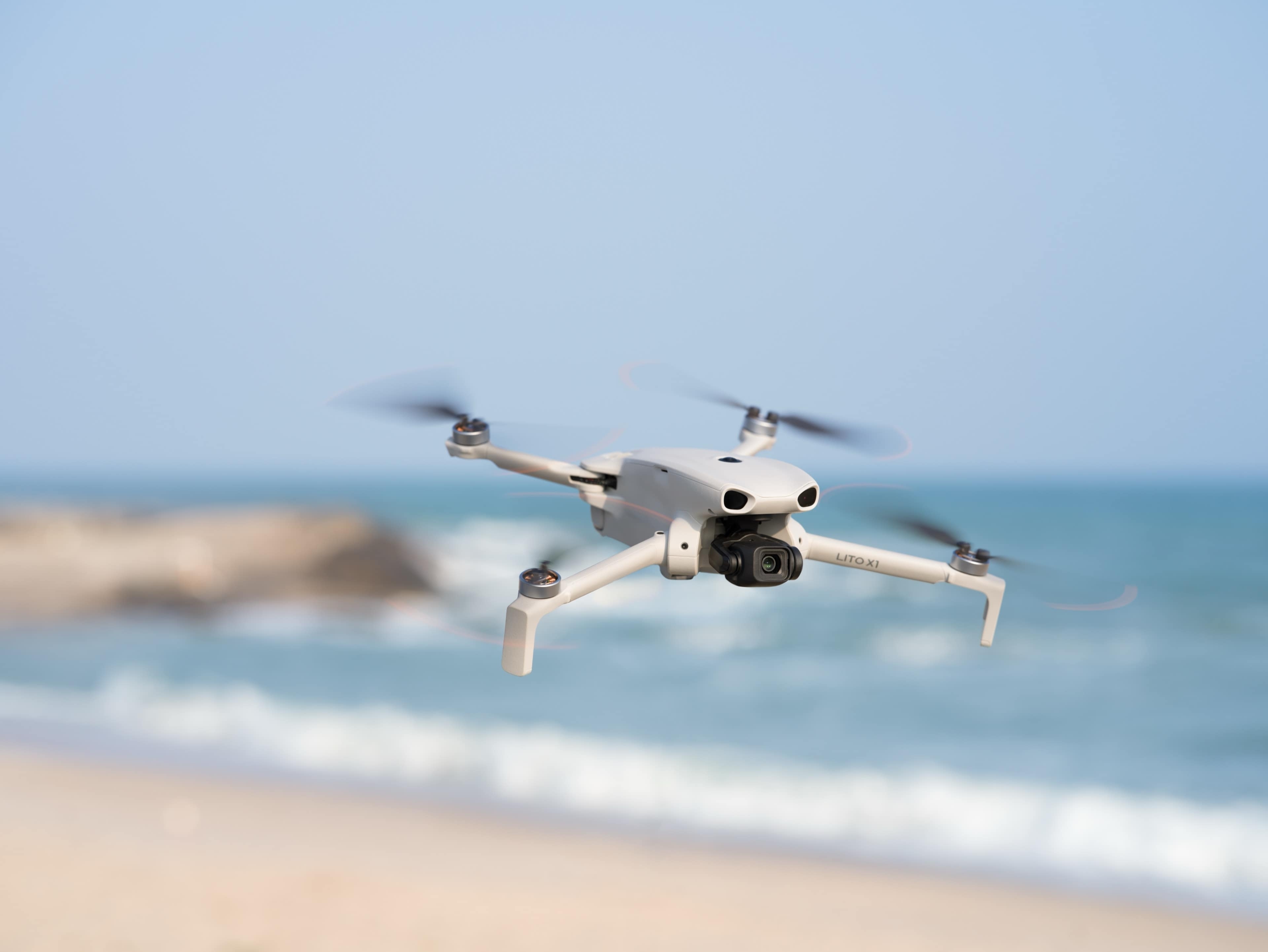 Fliegende DJI Lito X1 Fly More Combo (DJI RC 2) über Strand mit Wasser im Hintergrund.